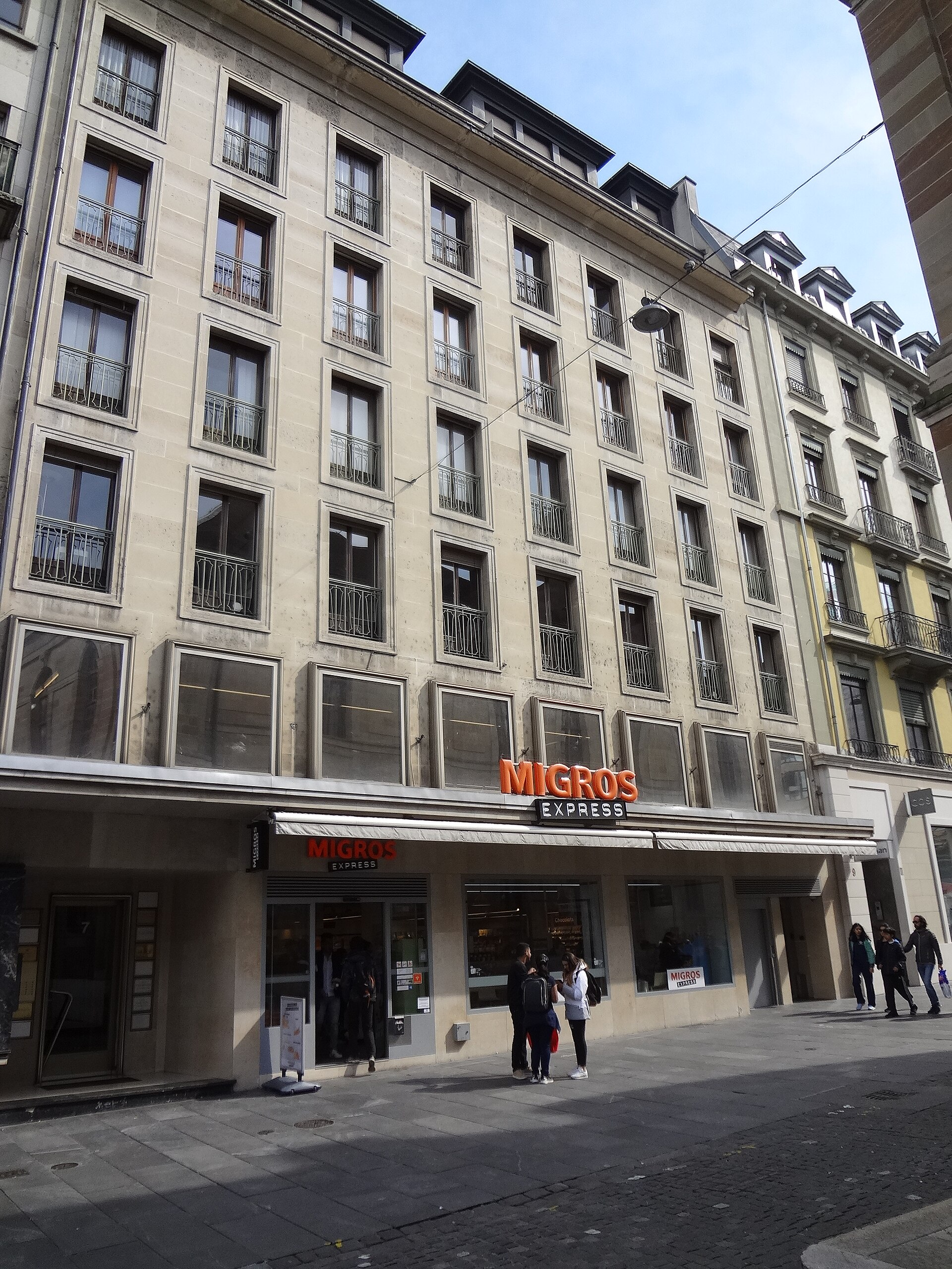 Migros Express signs mark a supermarket entrance on Place de la Fusterie in Geneva, with pedestrians beneath the tall stone facade.