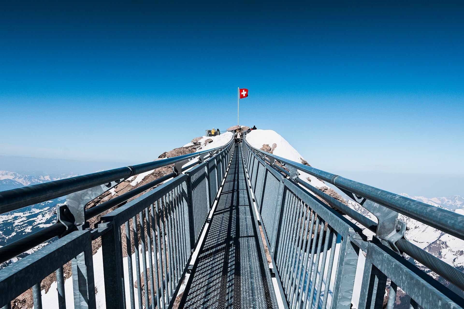 The narrow Peak Walk bridge leads toward a Swiss flag above snow-covered rocks at Glacier 3000 in clear weather.