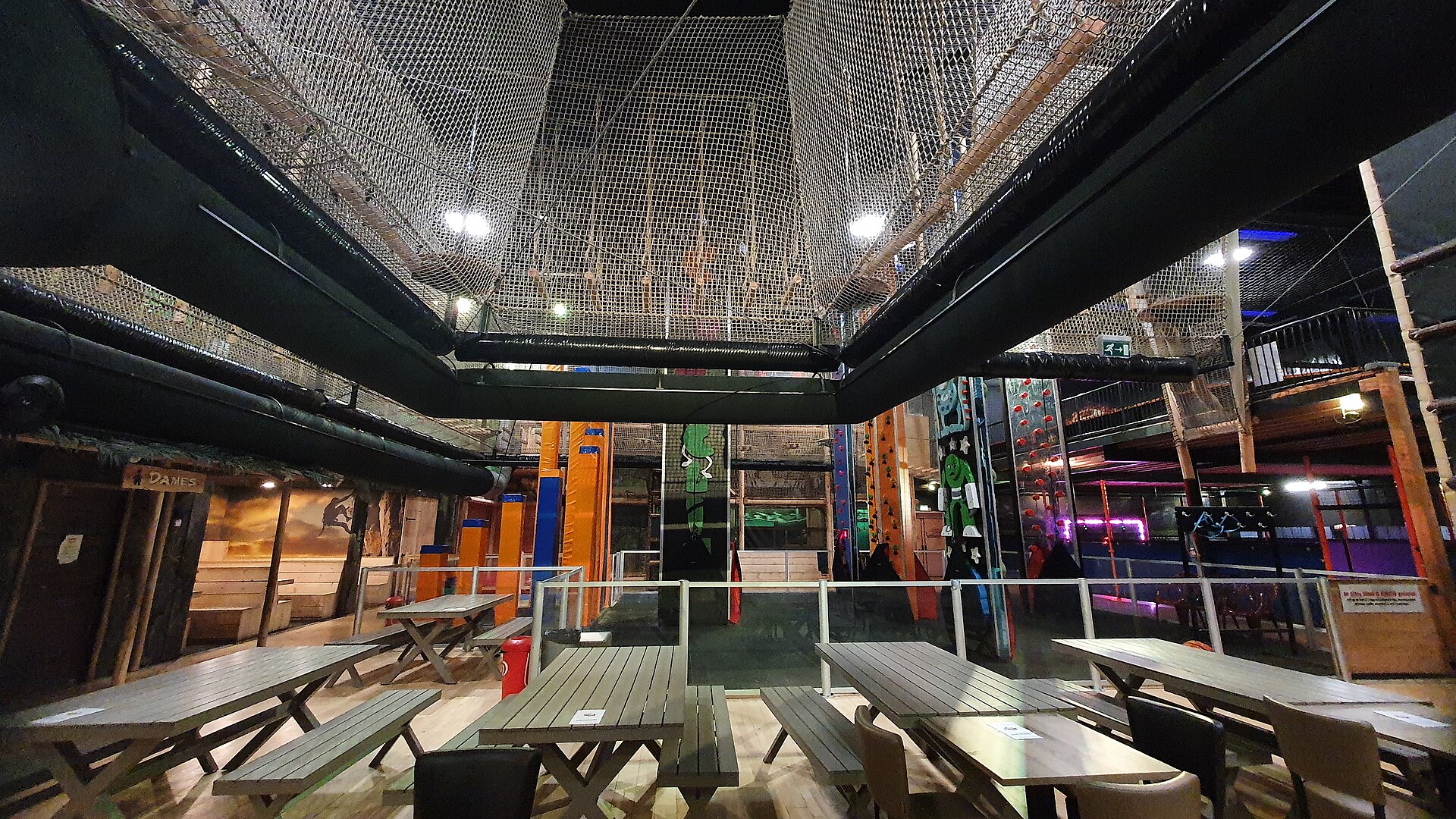 Netted play structures hang above wooden picnic tables beside colourful climbing walls at an indoor playground in the Netherlands.