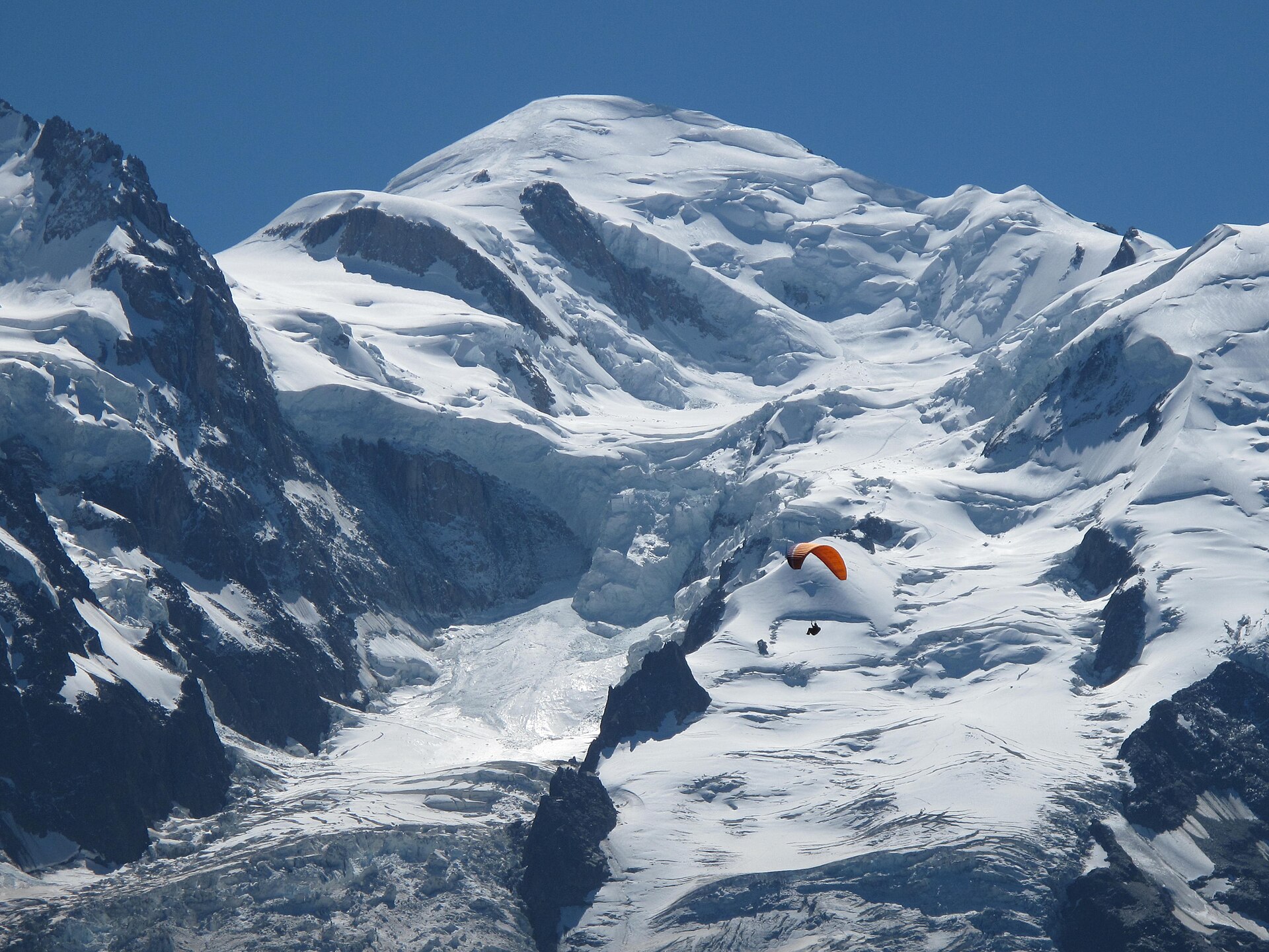 An orange paraglider crosses the snowy face of Mont Blanc, viewed from Planpraz on the Brevent side of Chamonix.