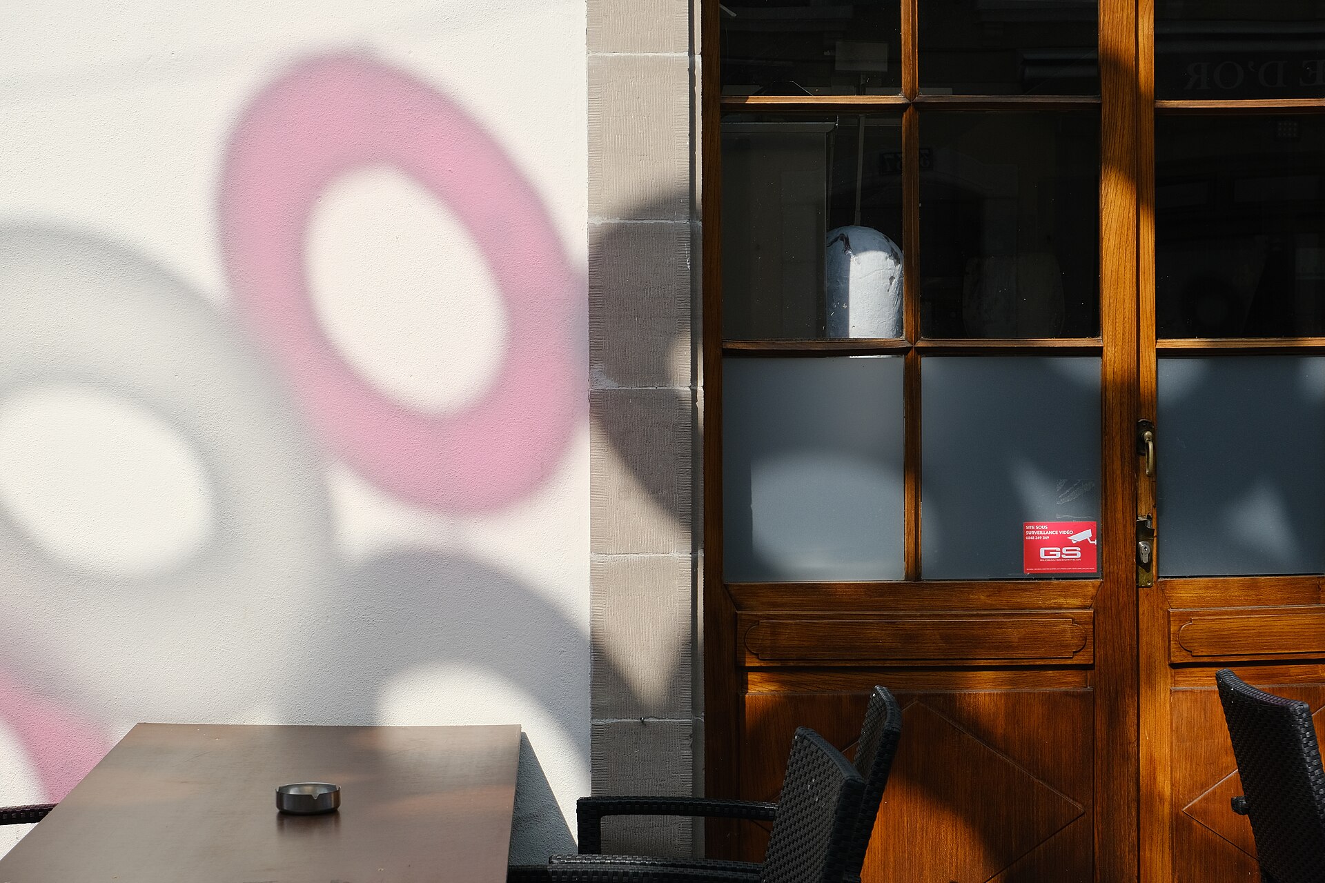 An outdoor cafe table and woven chairs stand beside a wooden doorway on Rue Saint-Joseph in Carouge, beneath colourful ring shadows.