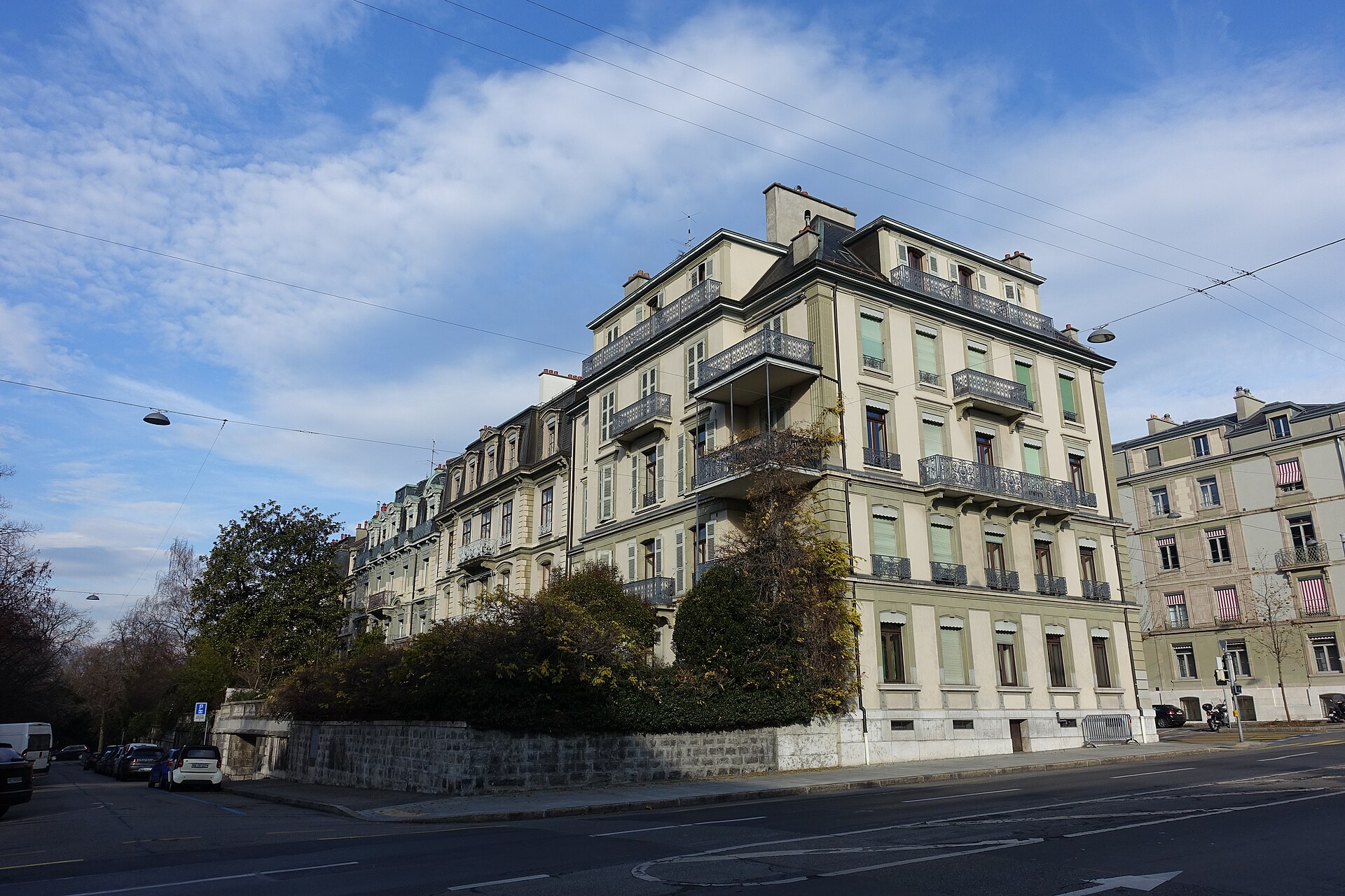 Pale residential buildings with green shutters and wrought iron balconies stand above a stone wall on a Geneva Champel street.