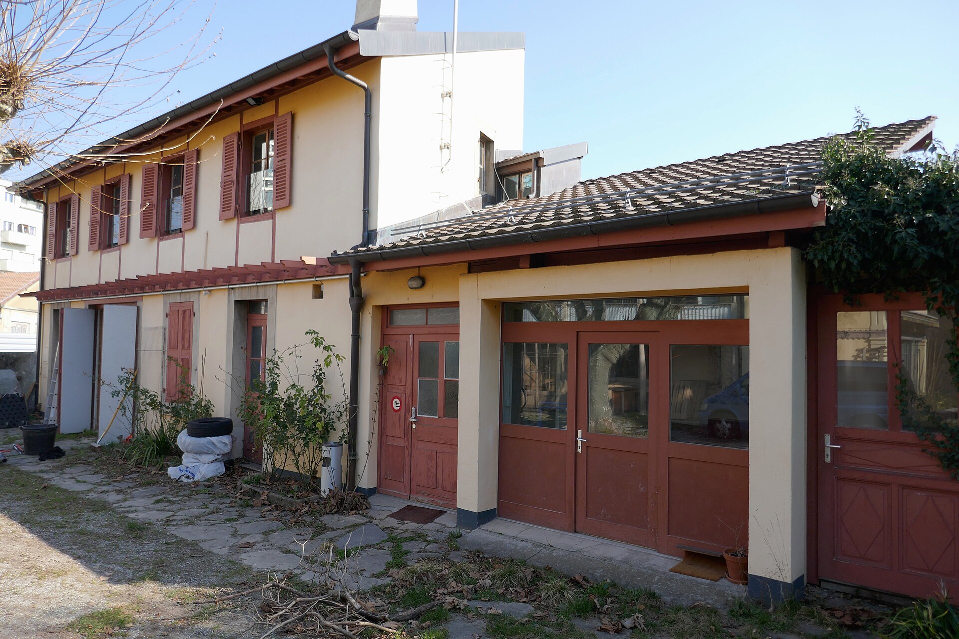 Pale yellow outbuildings with red-framed doors and shutters stand around a stone-paved, planted yard at Maison Baron in Carouge on a sunny day.
