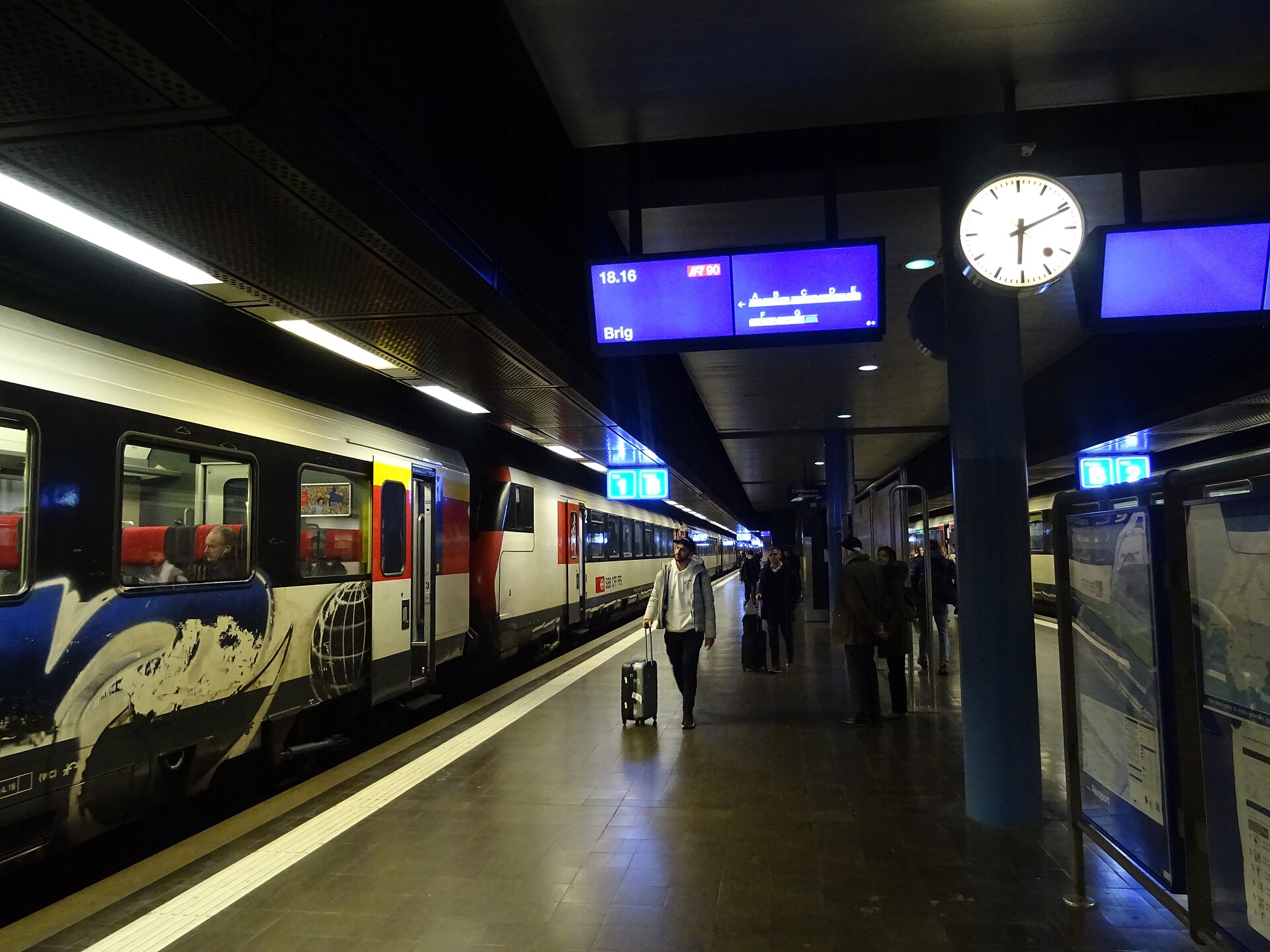 Passengers with luggage walk beside an InterRegio train at Geneva Airport station under a clock and a blue departure display.