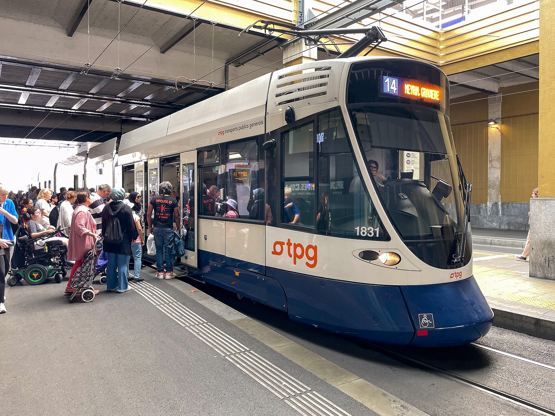 Passengers and a mobility scooter beside open doors of a blue and white TPG tram at Geneva Cornavin.