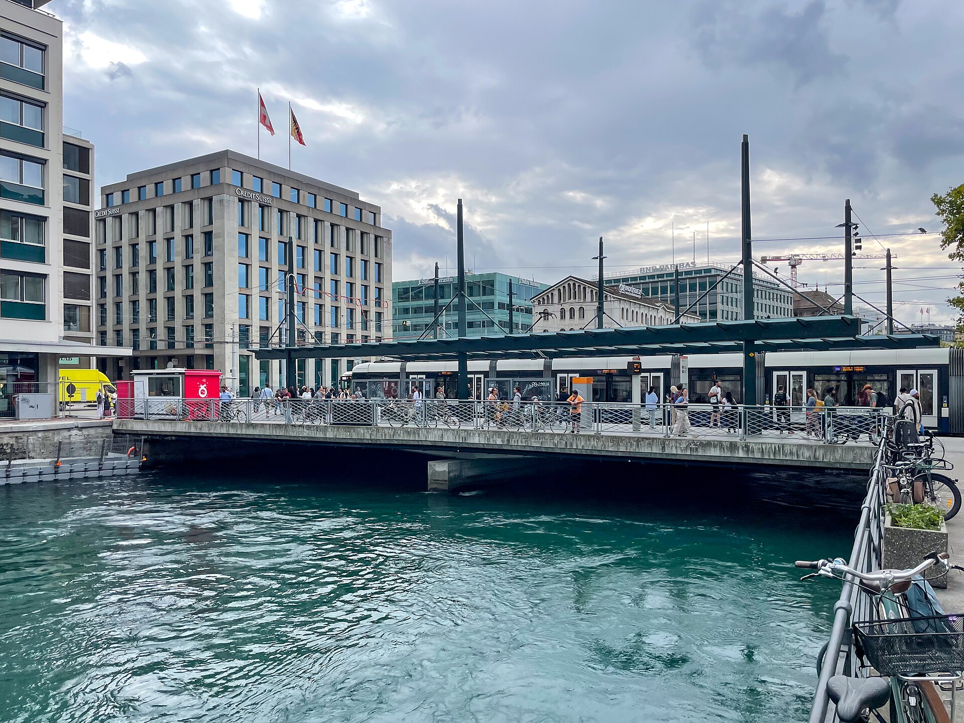 Passengers beside open tram doors at Ponts de l'Ile in Geneva, with bicycles and the Rhone in the foreground.