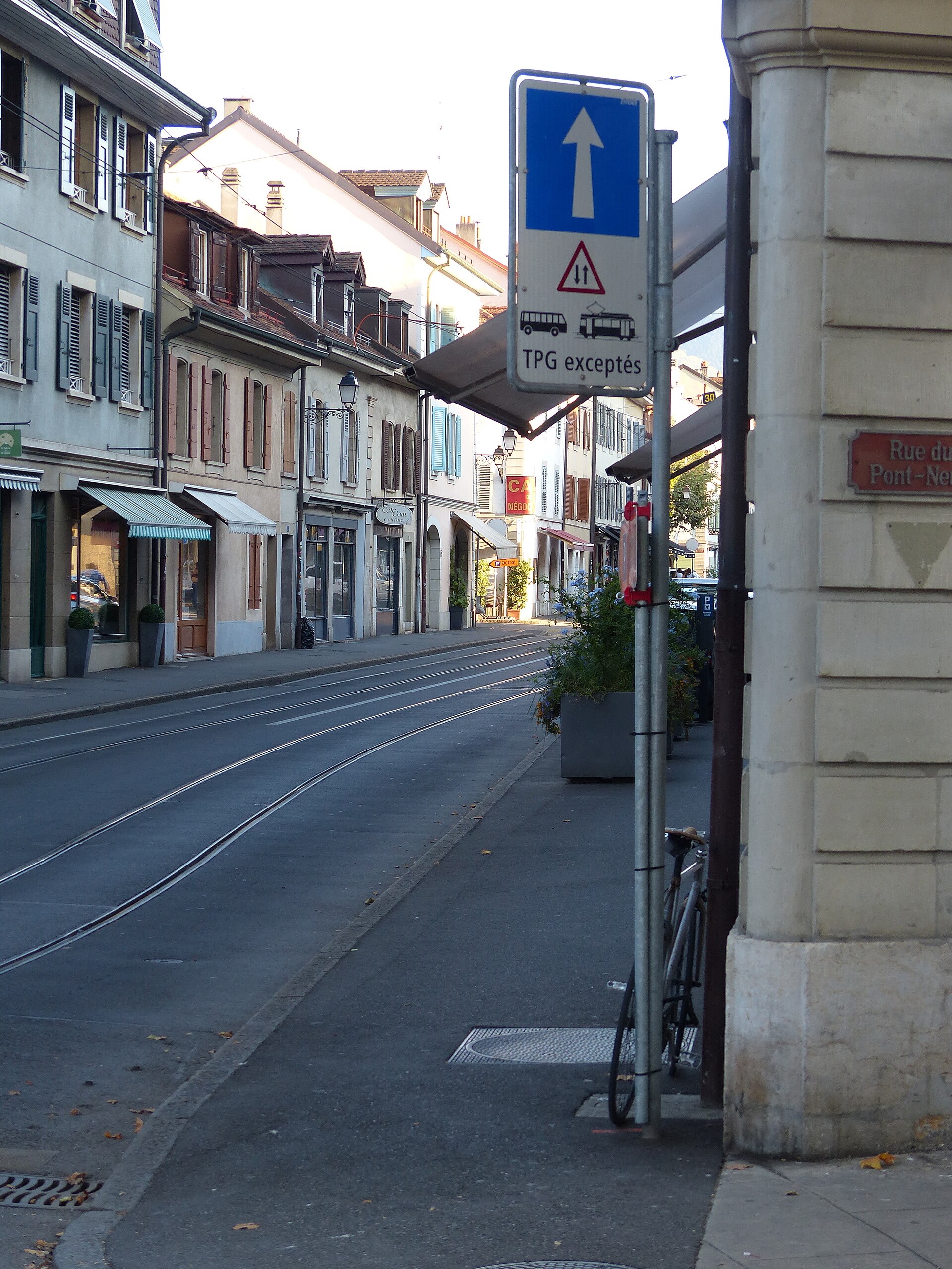 Pastel houses and striped shop awnings follow Carouge's tram tracks, with a Rue du Pont-Neuf plaque on the foreground corner.