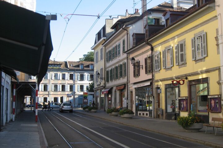 Pastel shuttered houses and ground-floor shops follow the curving tram tracks through Carouge on a sunny day.