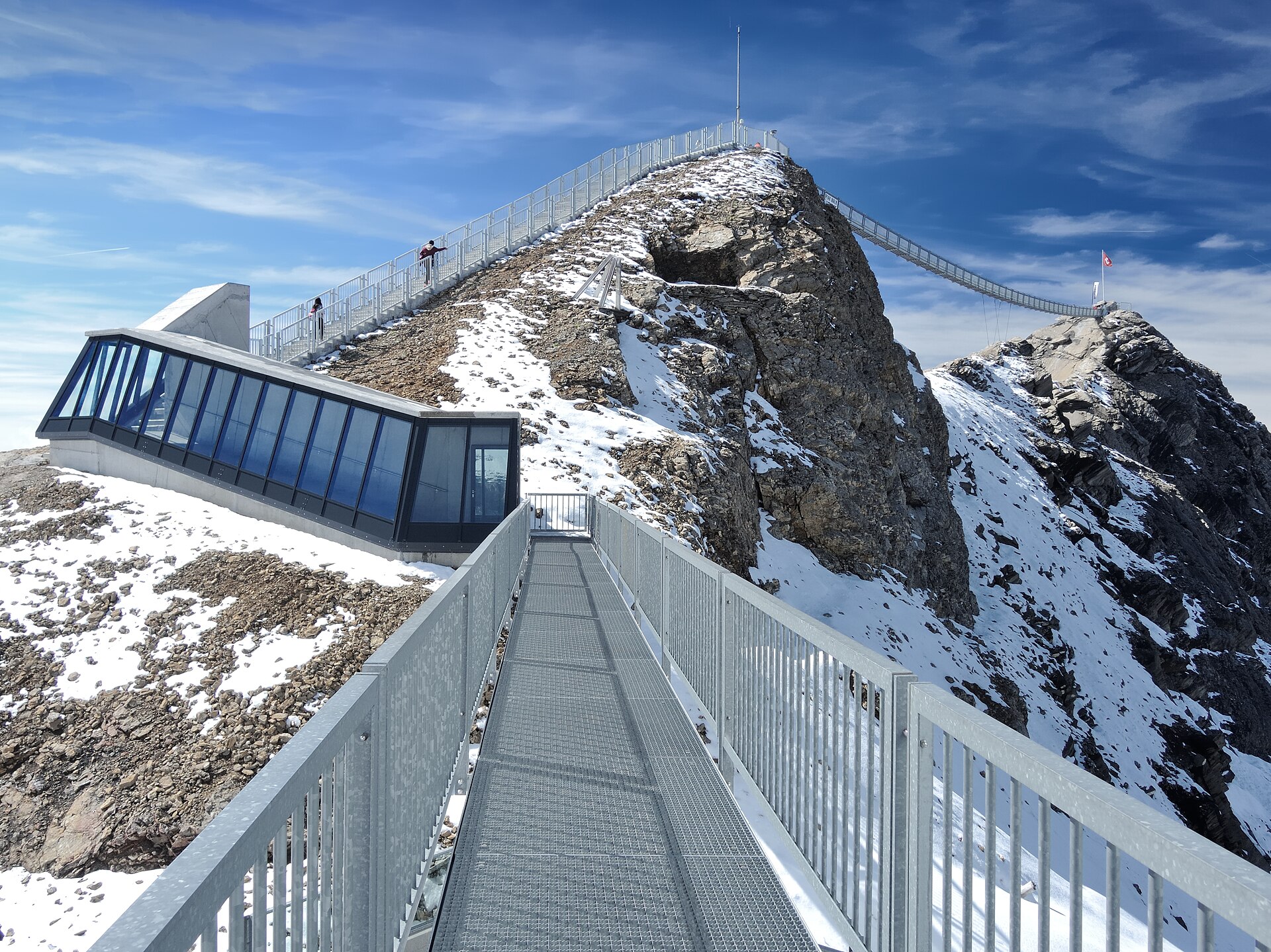 The Peak Walk suspension bridge spans two rocky summits at Glacier 3000 above metal walkways and patches of snow.