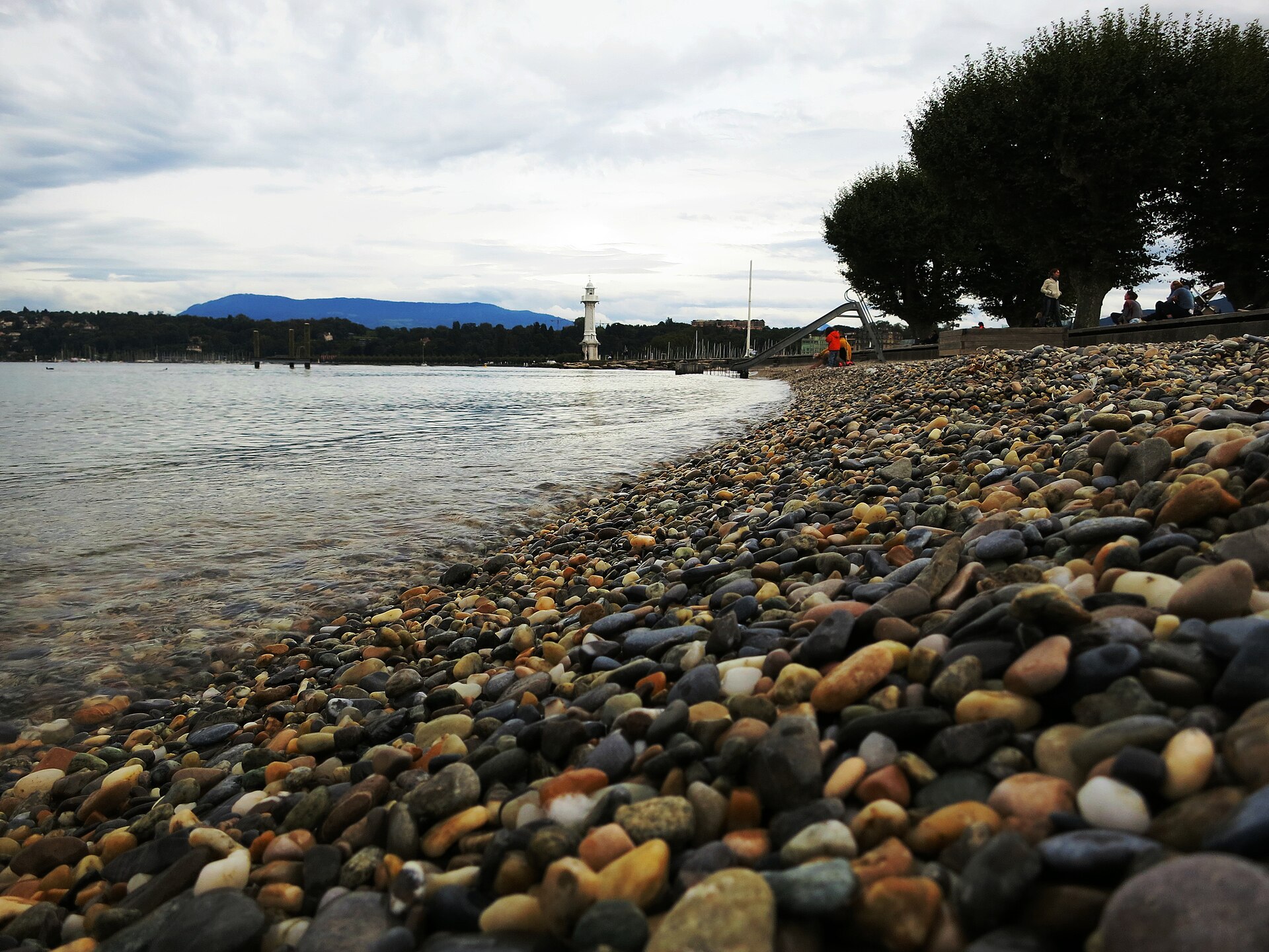 Pebbles on the shore at the Bains des Pâquis in Geneva, with calm lake water, a white lighthouse in the distance and plane trees on the right.