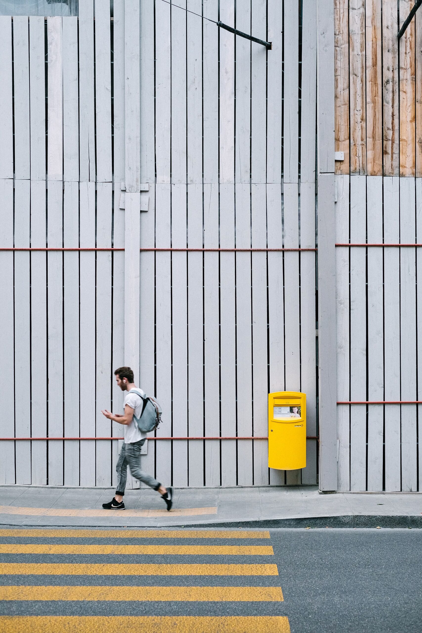 A pedestrian with a backpack walks past a yellow postbox and grey construction hoarding beside a Geneva street crossing.