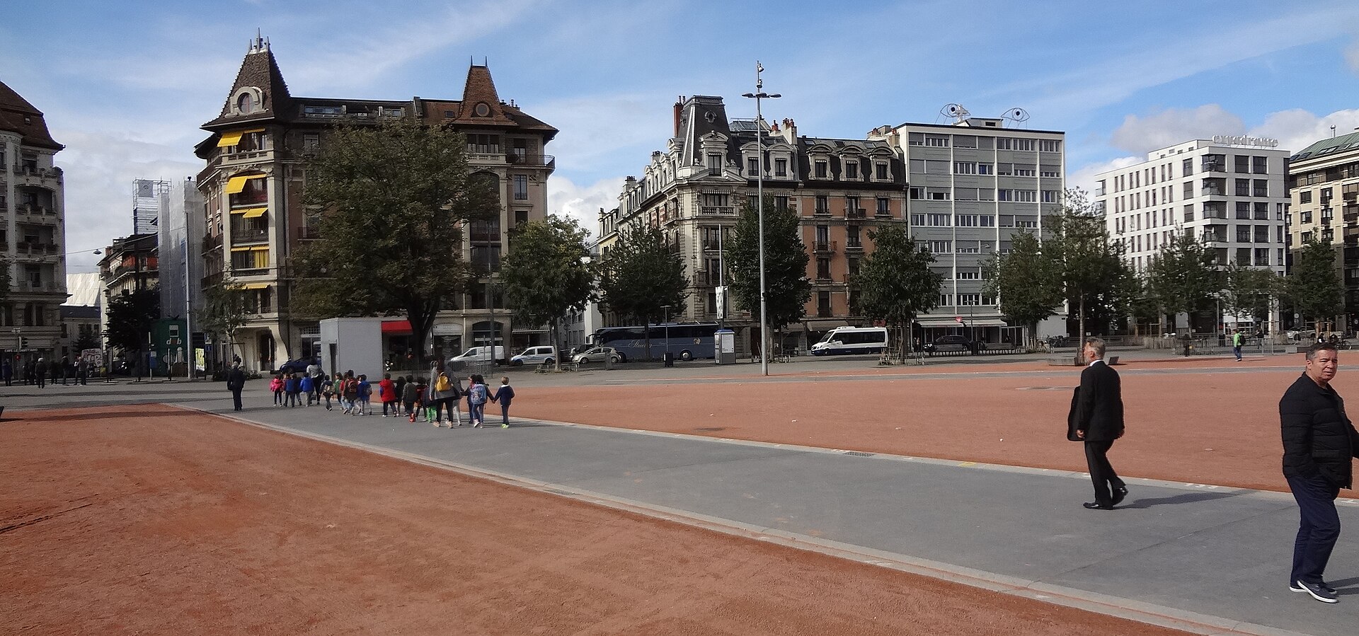 Pedestrians cross paved paths on Geneva's red Plaine de Plainpalais, with ornate corner buildings and trees along its edge.