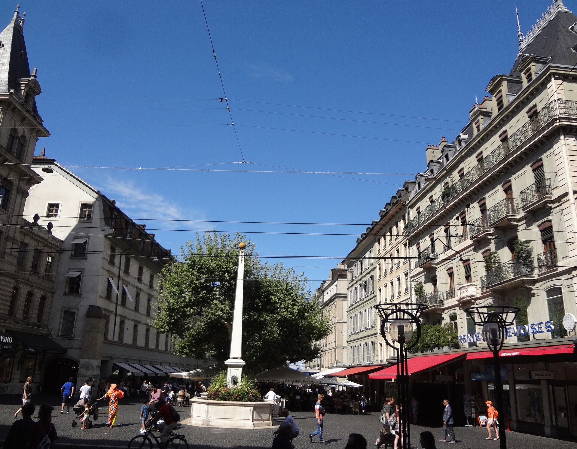 People cross Geneva's Place du Molard beside a flower-planted fountain, cafe terraces and rows of stone buildings.