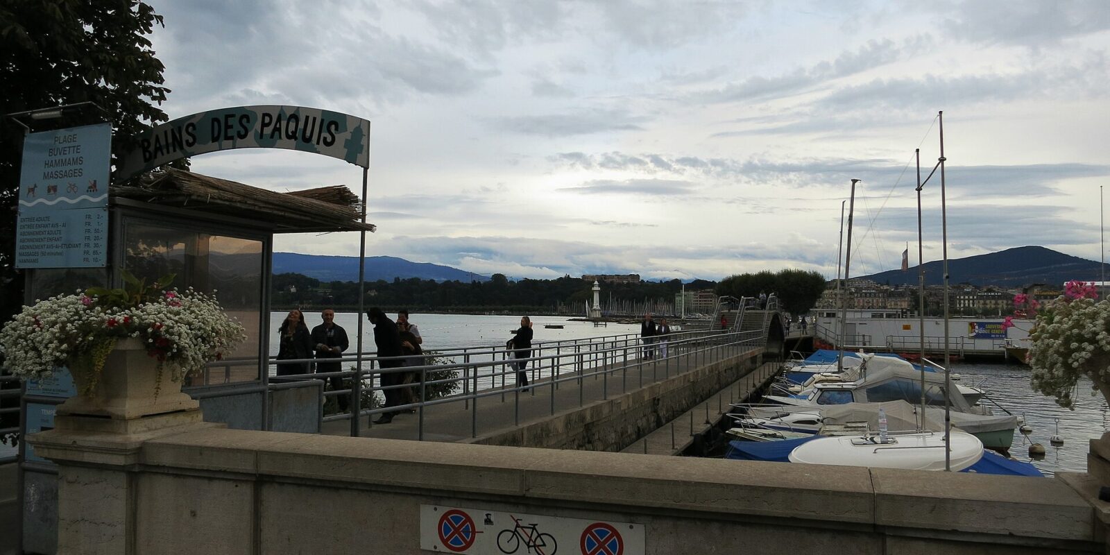 People in jackets walk beside moored boats at Geneva's Bains des Pâquis entrance beneath a layered grey cloud sky.