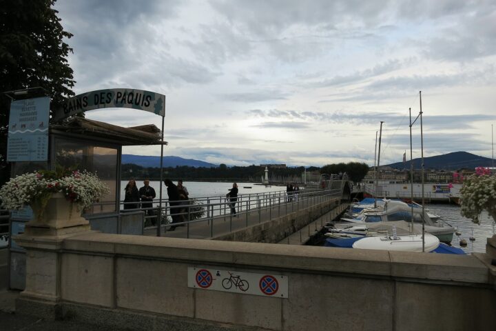 People in jackets walk beside moored boats at Geneva's Bains des Pâquis entrance beneath a layered grey cloud sky.