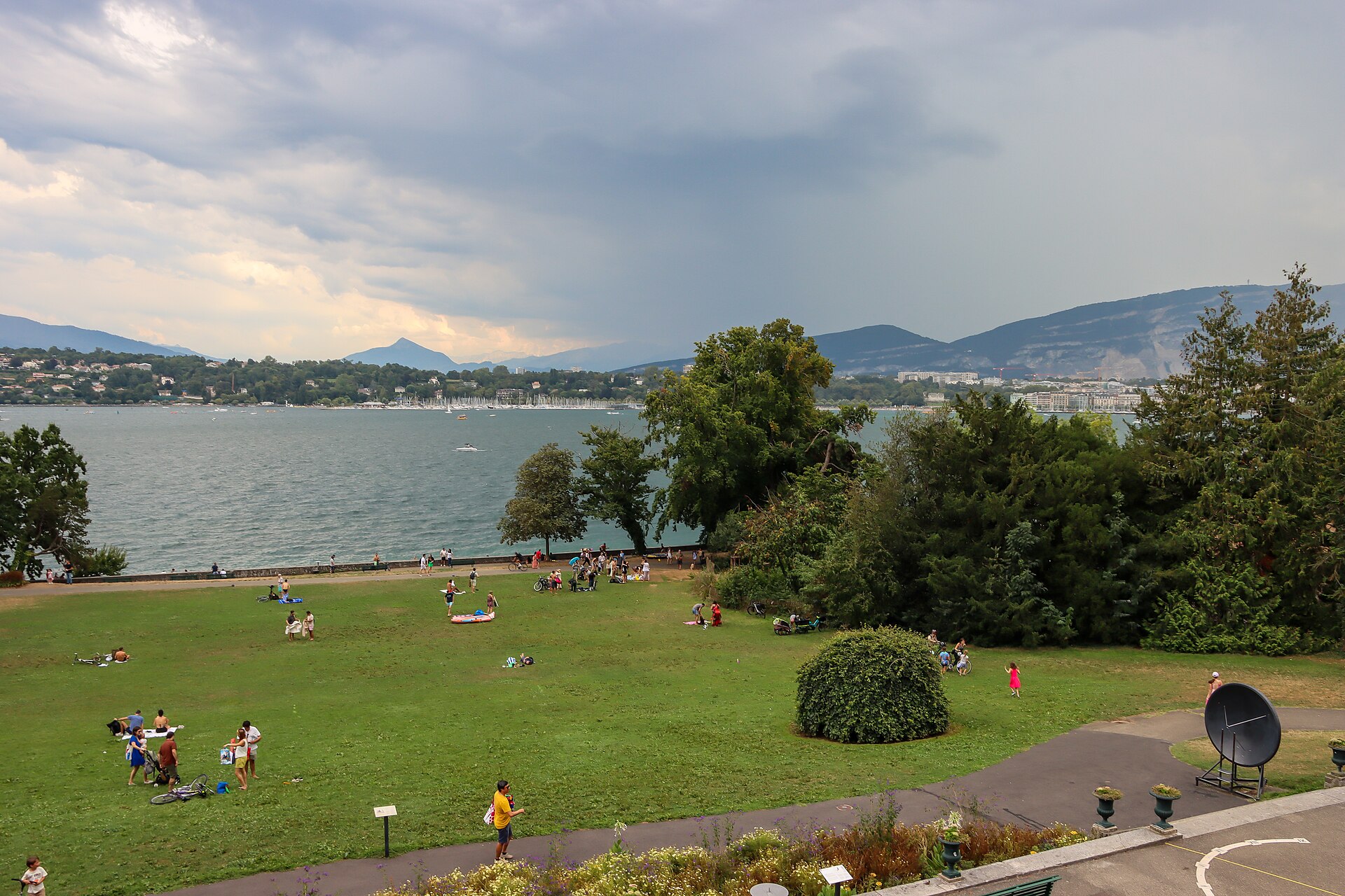 People in light summer clothing gather on Geneva’s Perle du Lac lawn beside the lake under cloudy skies.