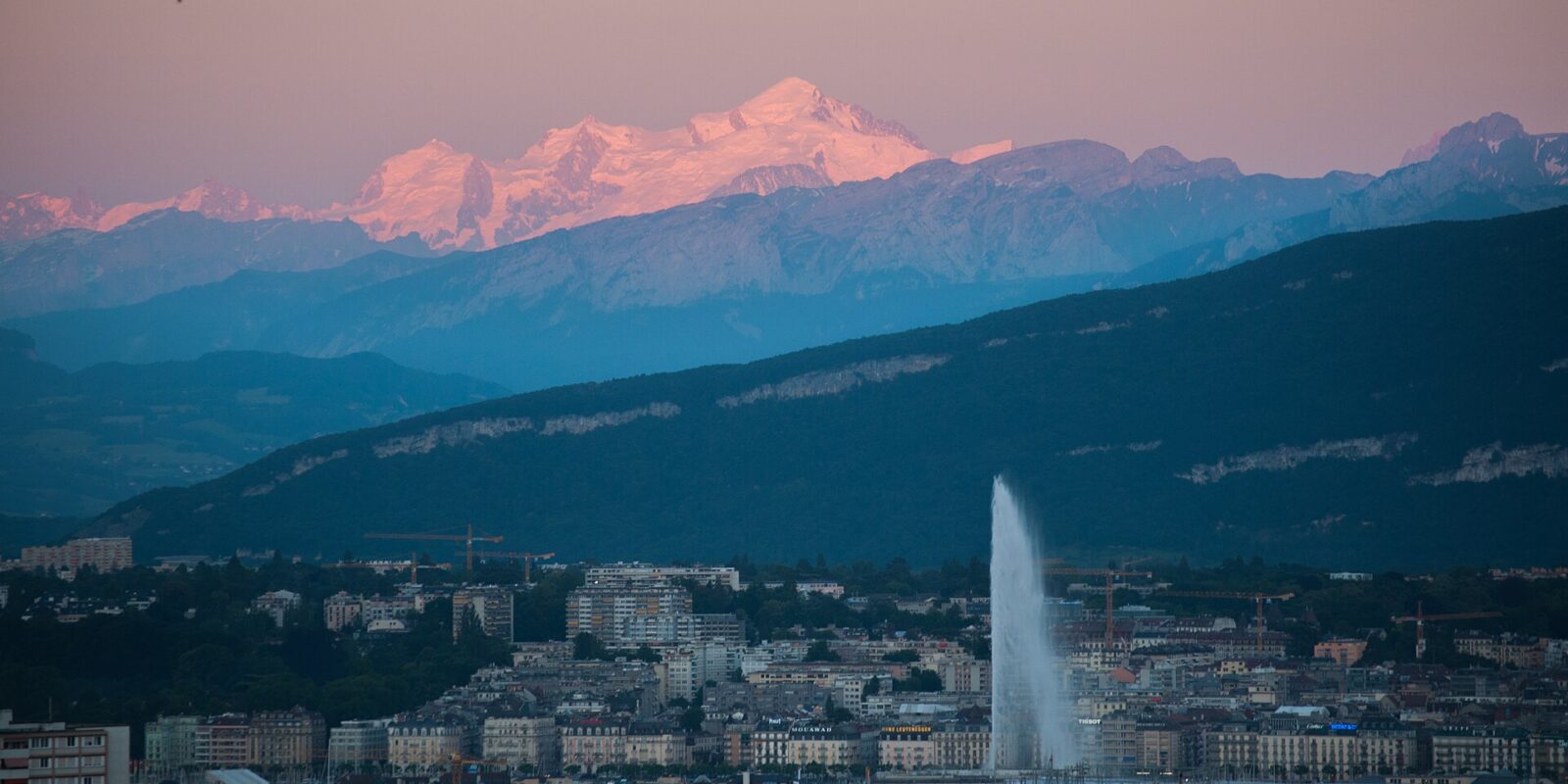 Pink evening light touches Mont Blanc above Geneva's waterfront buildings and the Jet d'Eau, with layered mountain ridges between.