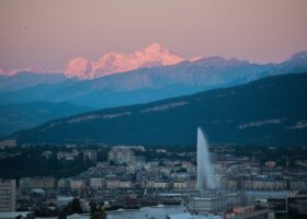 Pink evening light touches Mont Blanc above Geneva's waterfront buildings and the Jet d'Eau, with layered mountain ridges between.