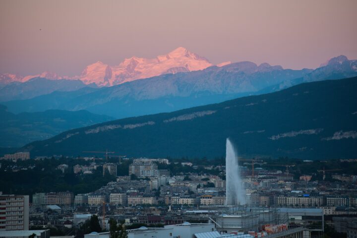 Pink evening light touches Mont Blanc above Geneva's waterfront buildings and the Jet d'Eau, with layered mountain ridges between.