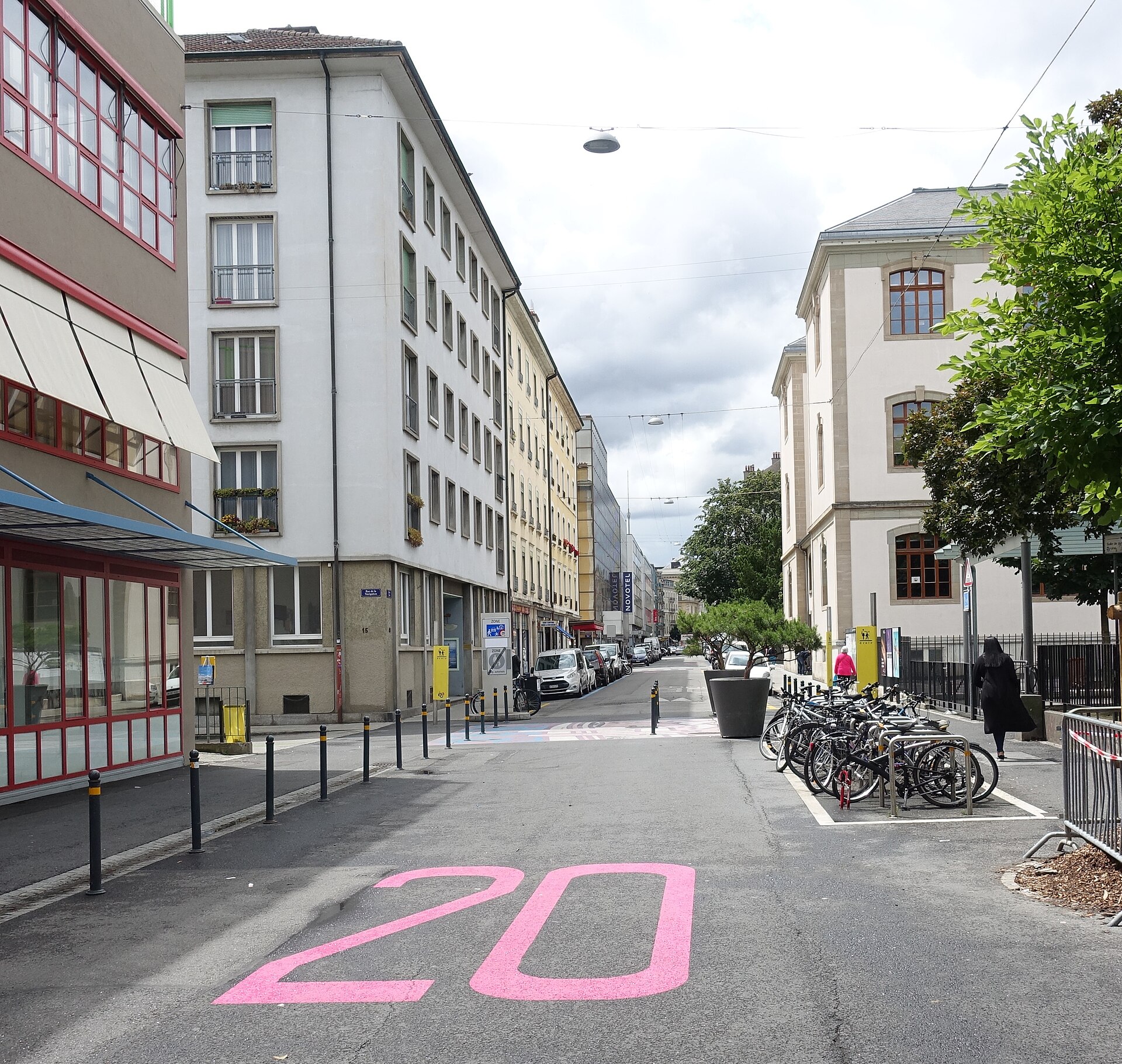 A pink speed marking, parked bicycles and pedestrians fill Rue de Berne in Geneva's Pâquis neighborhood beneath apartment buildings.