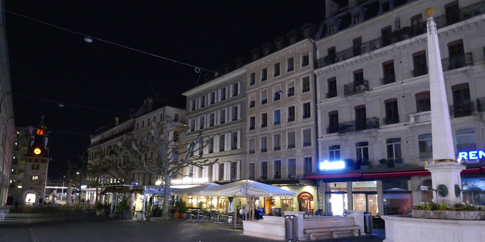 Place du Molard in Geneva at night, with restaurant terraces under white parasols below stone apartment buildings, the Molard column at right and the clock tower lit at left