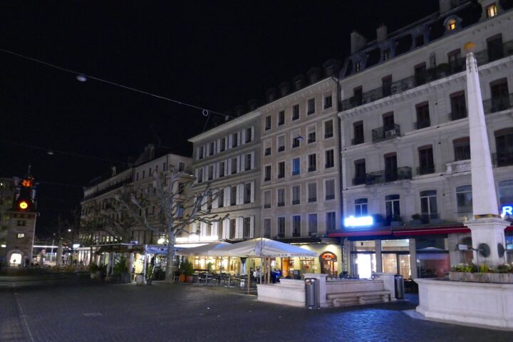 Place du Molard in Geneva at night, with restaurant terraces under white parasols below stone apartment buildings, the Molard column at right and the clock tower lit at left