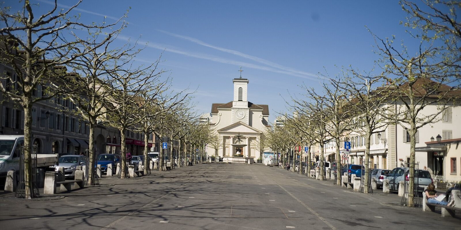 Pollarded trees and low shuttered houses frame Place du Marche in Carouge, with Sainte-Croix church at the far end.