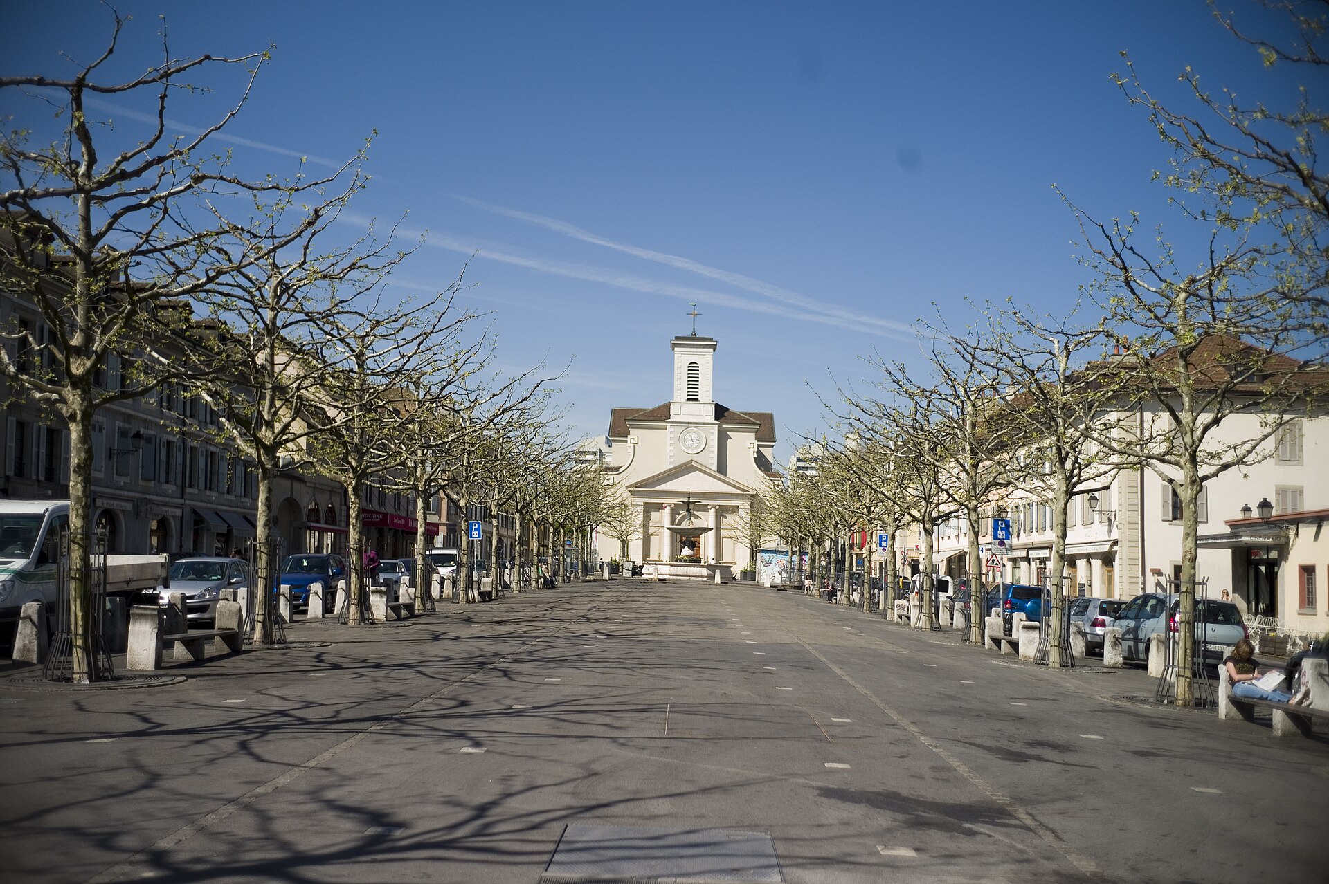 Pollarded trees and low shuttered houses frame Place du Marché in Carouge, with Sainte-Croix church at the far end.