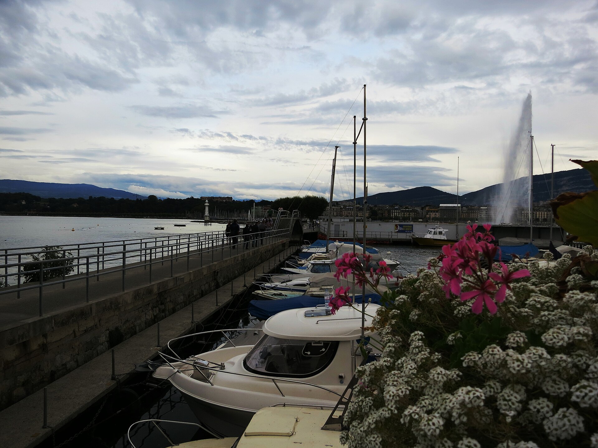A railed walkway slopes along Geneva's Pâquis jetty beside moored boats, with pedestrians and the Jet d'Eau beyond.
