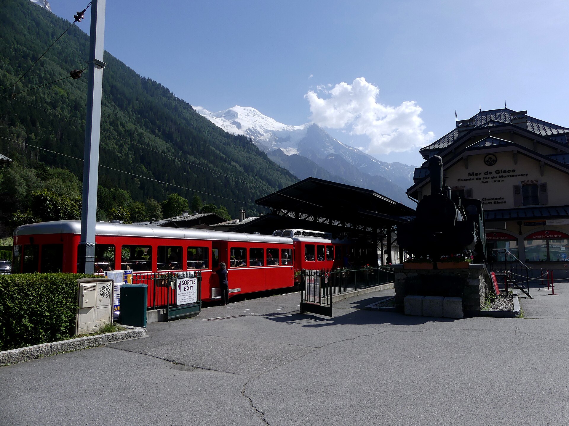 A red Montenvers train waits beside Chamonix’s Mer de Glace station, with a preserved locomotive and snowy peaks behind.