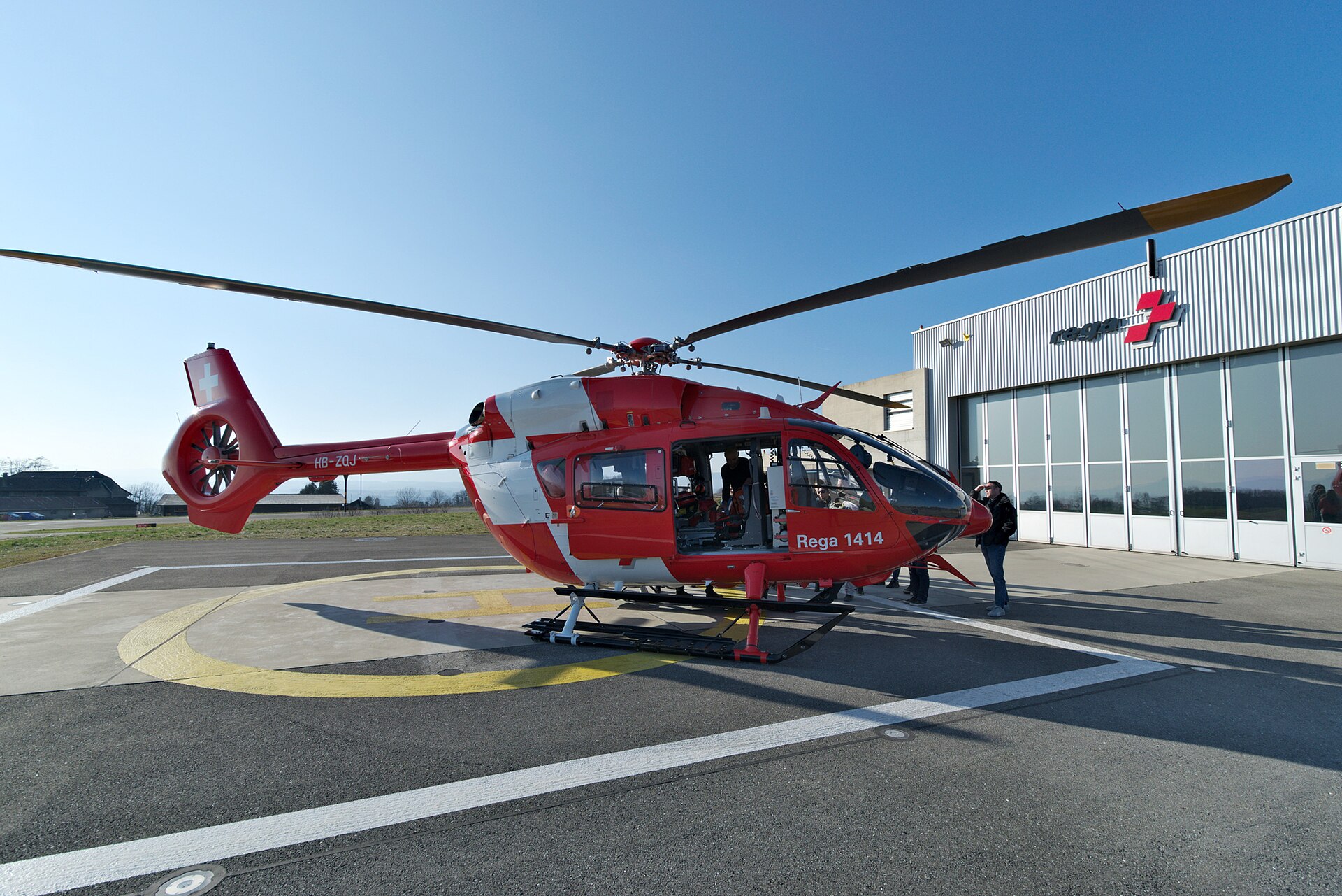 A red and white Rega rescue helicopter stands outside its hangar at Lausanne Airport, with its side door open.