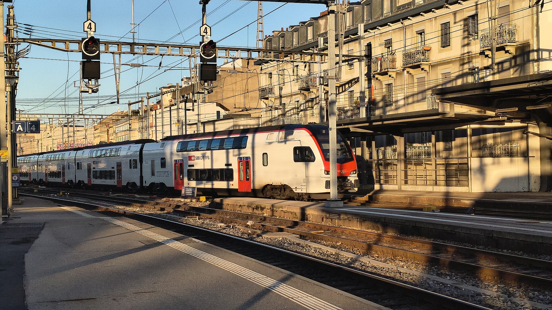 A red-and-white SBB double-deck train arrives at Geneva Cornavin beside sunlit buildings, overhead wires and railway signals.