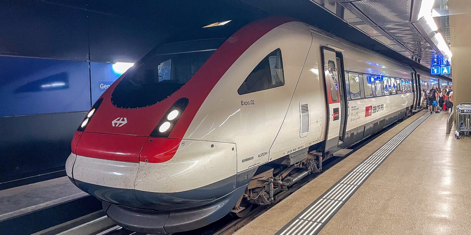 A red-and-white SBB train waits at Geneva Airport’s underground station, with passengers and luggage farther along the platform.