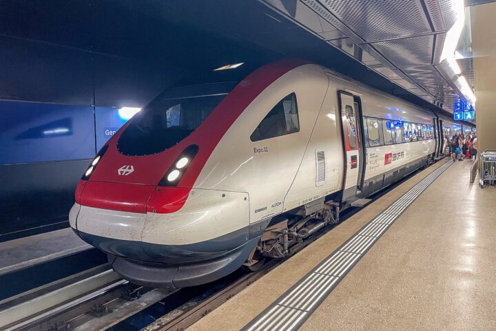 A red-and-white SBB train waits at Geneva Airport’s underground station, with passengers and luggage farther along the platform.