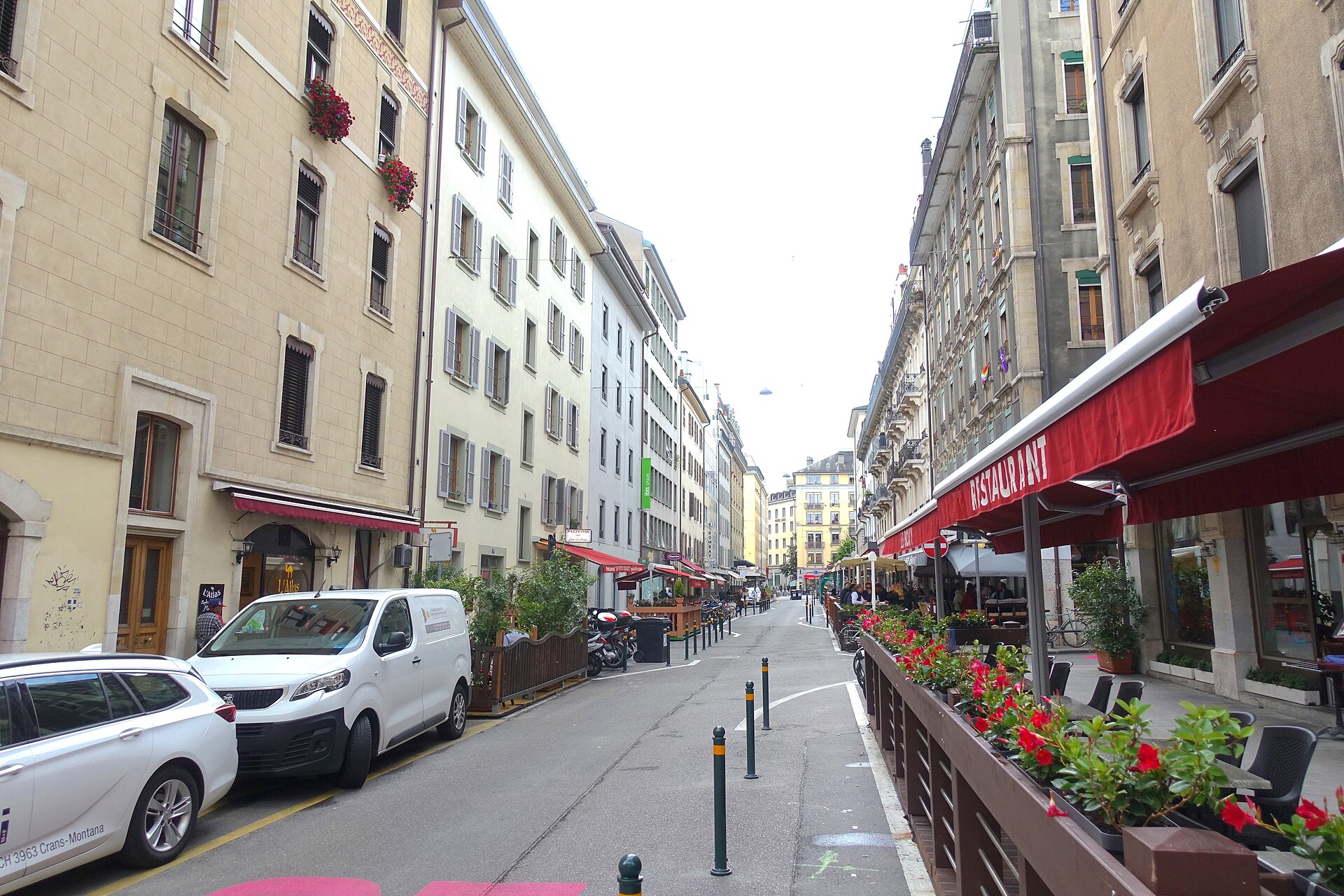 Restaurant terraces with red awnings and flower-filled barriers line Rue Chaponnière in Geneva, with tables beside the narrow street.