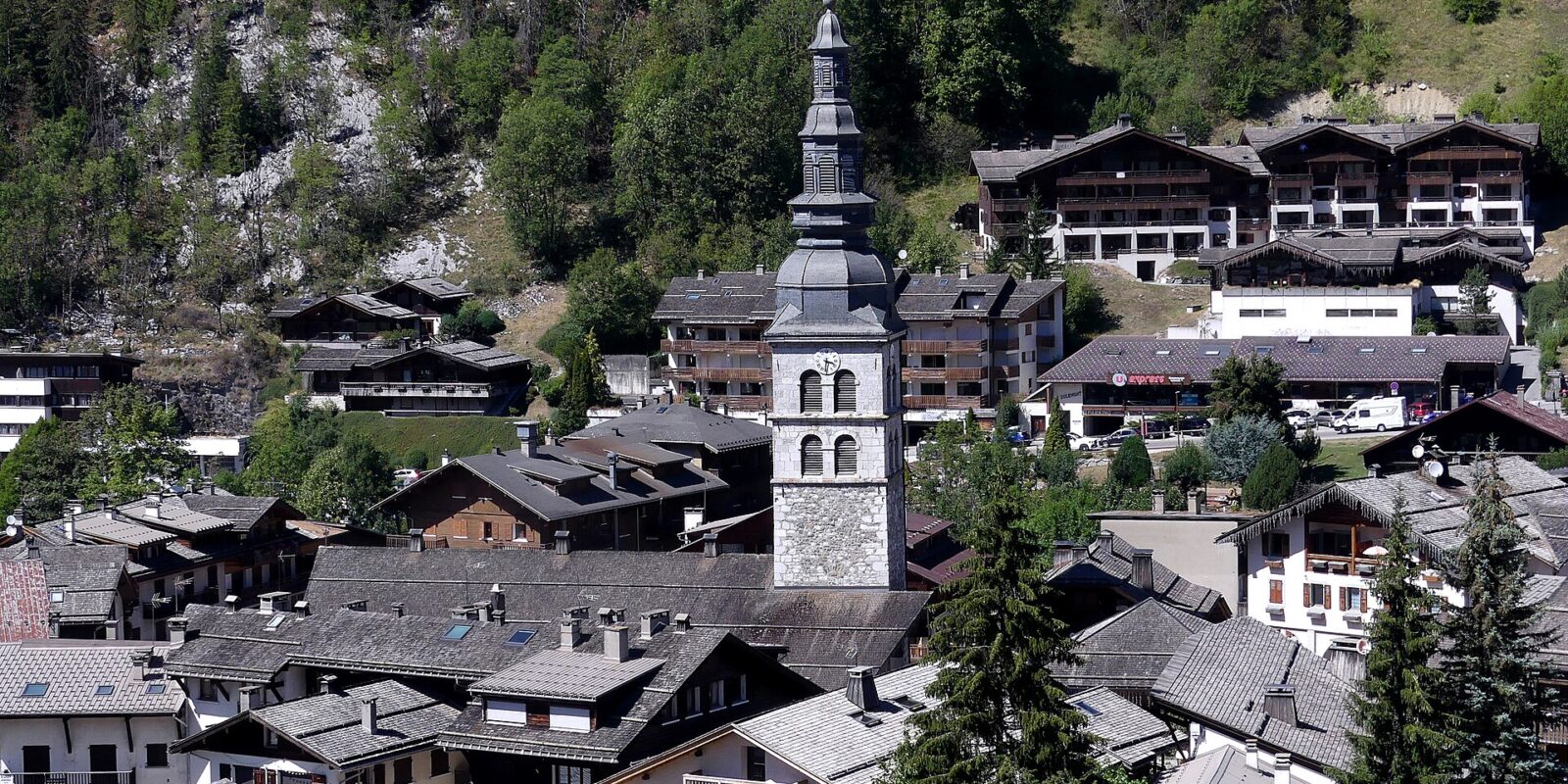 Sainte-Foy church rises among chalet rooftops in La Clusaz, backed by a wooded hillside in summer sunshine.