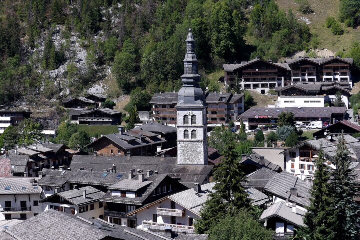 Sainte-Foy church rises among chalet rooftops in La Clusaz, backed by a wooded hillside in summer sunshine.
