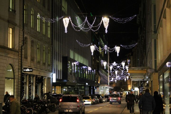 Seasonal lights hang above Geneva's Rue du Rhône, with Vacheron Constantin signage, pedestrians and evening traffic beneath illuminated shopfronts.