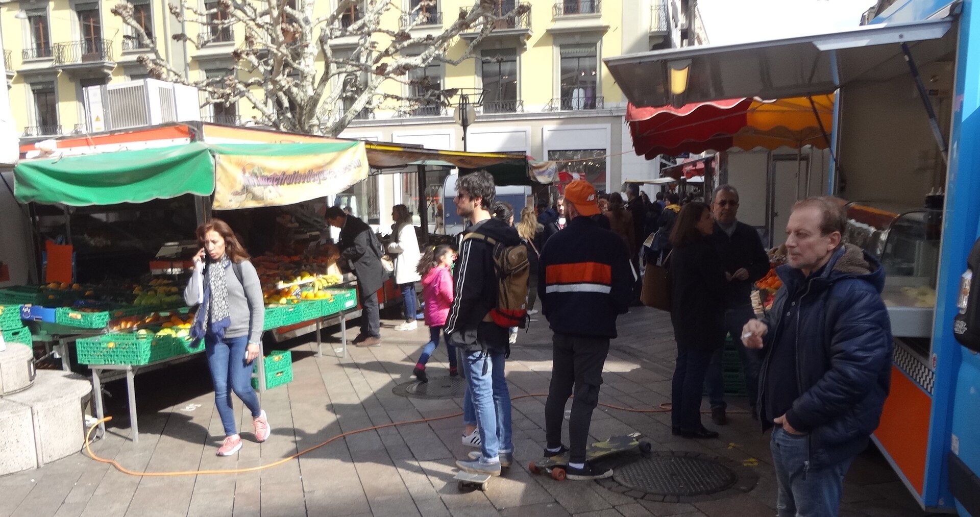 Shoppers browse fruit and vegetable stalls and a food truck at the Saturday market on Place de la Fusterie in Geneva, with apartment buildings behind.