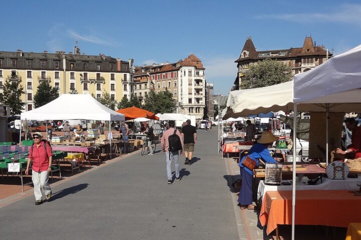 Shoppers pass book tables and secondhand stalls beneath white awnings on the red Plainpalais square in Geneva.