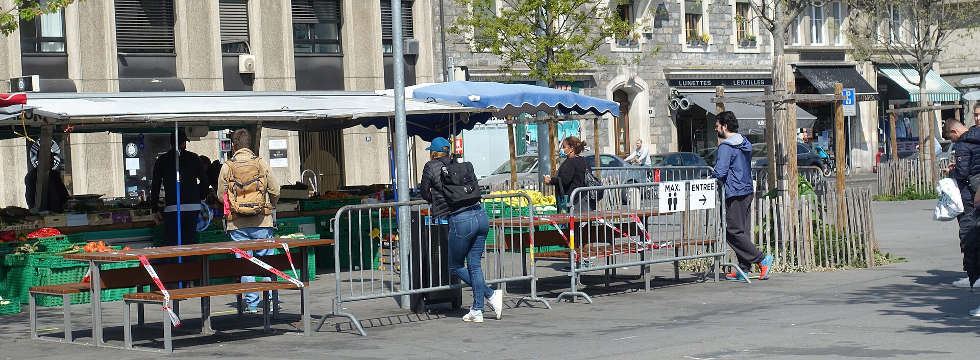 Shoppers queue beside green produce crates beneath blue and white canopies at Geneva's Plainpalais market in April 2020.