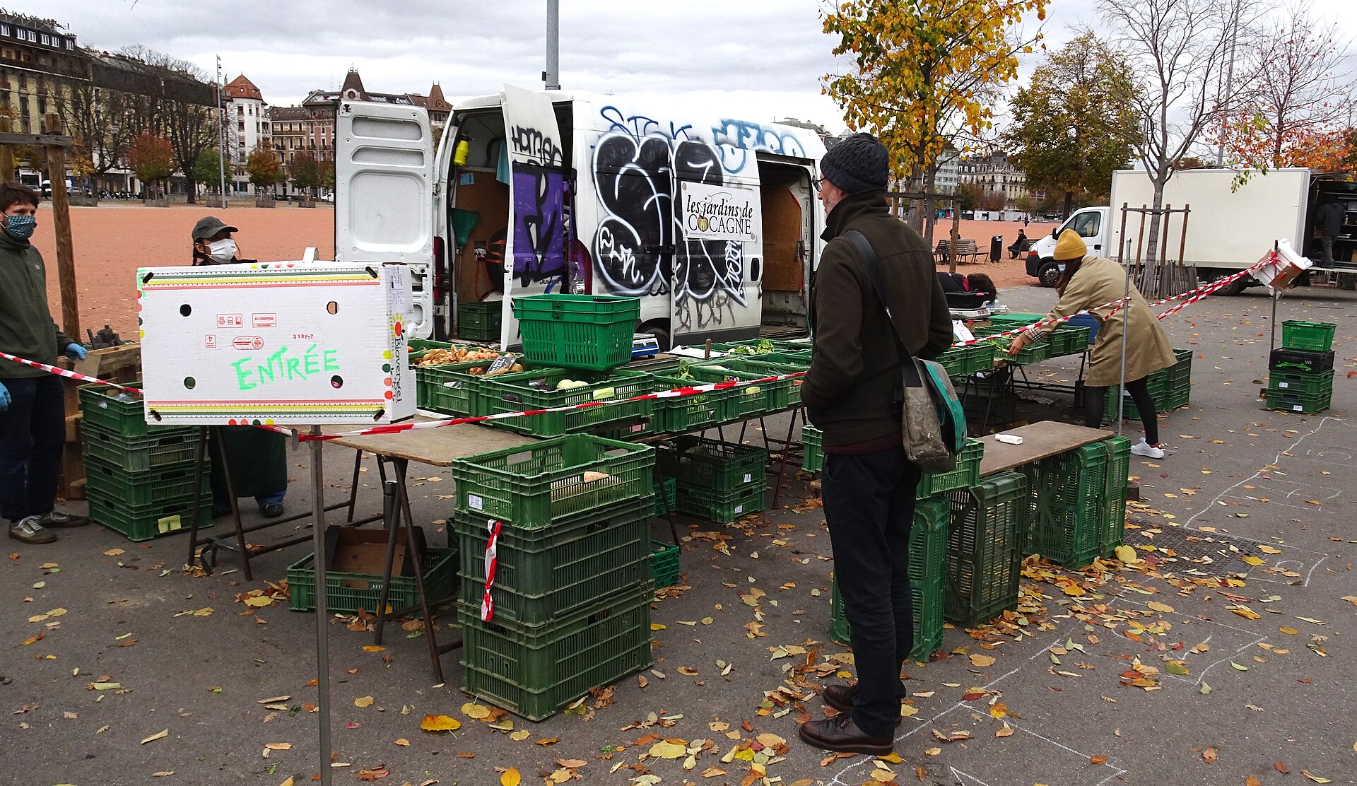 Shoppers visit a vegetable stall beside the red gravel plain at Plainpalais market in Geneva on an autumn day.
