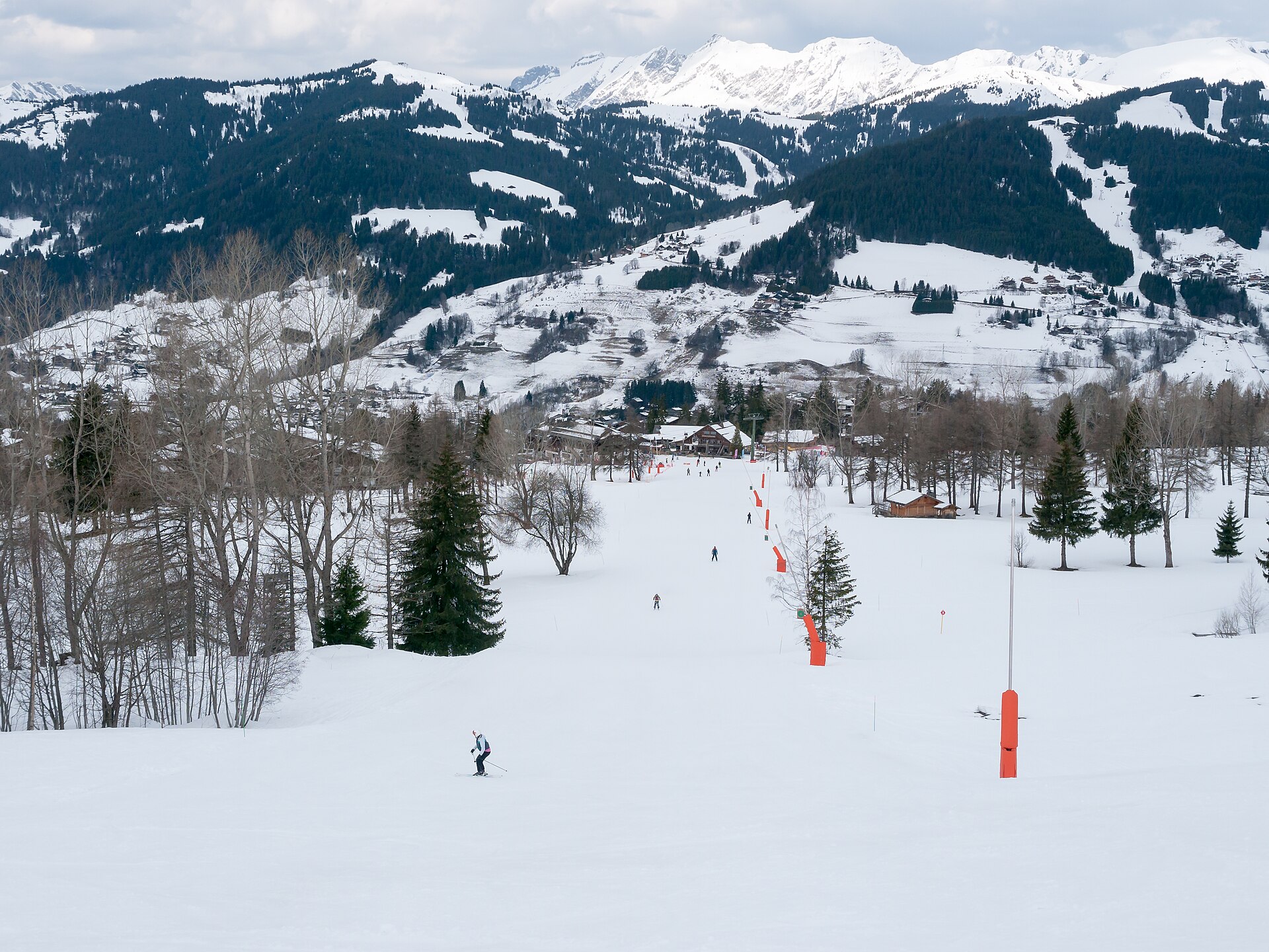 Skiers descend a broad tree-lined piste in the Evasion Mont-Blanc ski area at Saint-Gervais, with chalets and snowy ridges beyond.