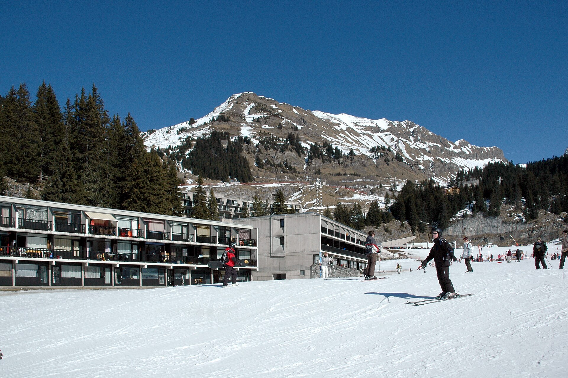 Skiers descend past the buildings of Flaine Front de Neige beneath a rocky, partly snow-covered mountain.