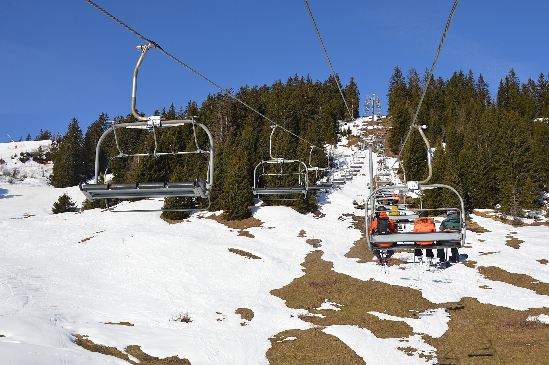 Skiers ride the Ravine chairlift above patchy snow and exposed grass between conifers in the Le Jaillet ski area near Megève.
