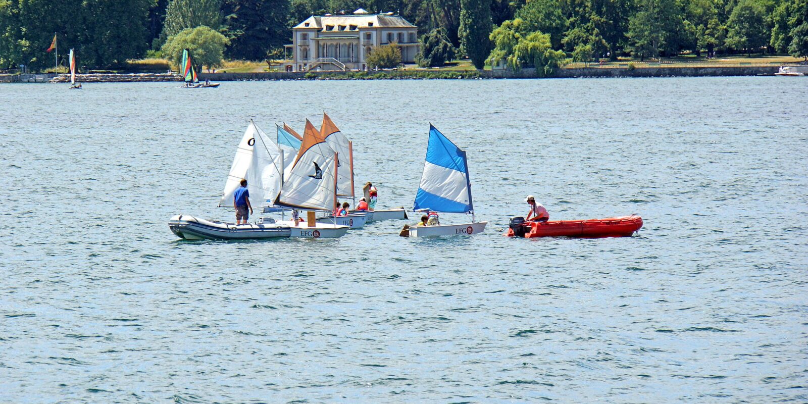 Small sailing dinghies gather beside a red support boat on Lake Geneva, with Geneva's tree-lined waterfront and buildings behind them.