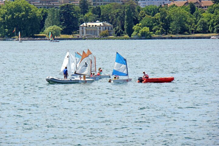 Small sailing dinghies gather beside a red support boat on Lake Geneva, with Geneva's tree-lined waterfront and buildings behind them.