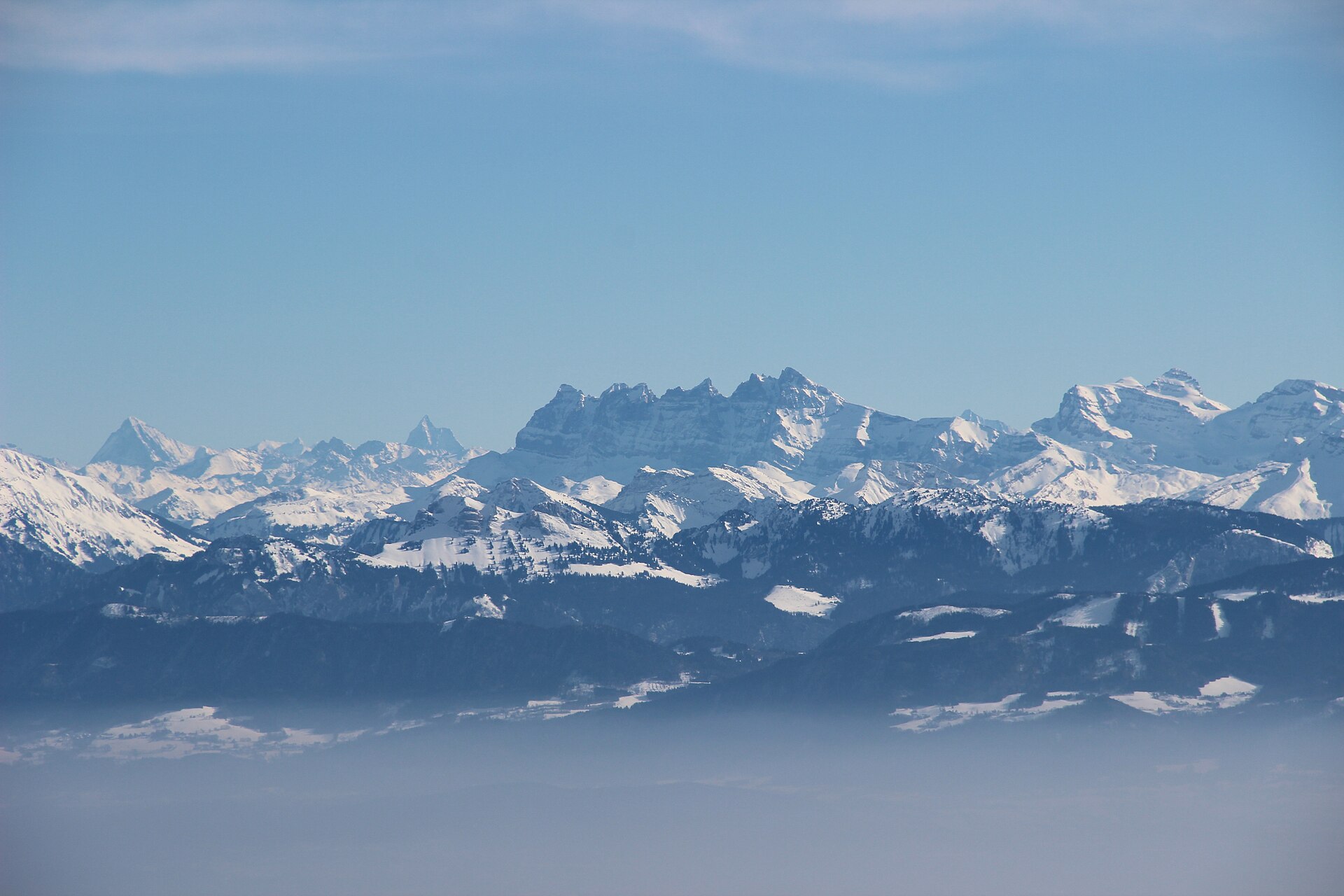 Snow-covered Alpine peaks including the Dents du Midi rise under a blue sky above a band of grey-blue fog that fills the lowlands, seen from La Dôle