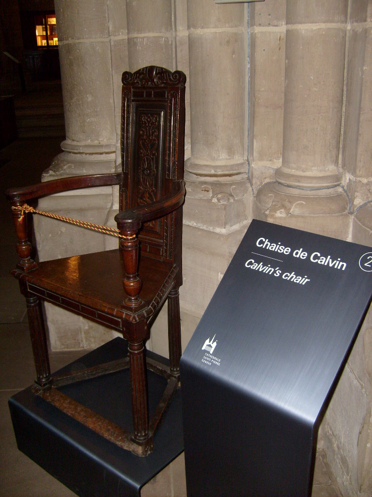 A dark wooden high-backed chair with carved tracery in St Pierre Cathedral, beside a sign reading Chaise de Calvin, Calvin's chair