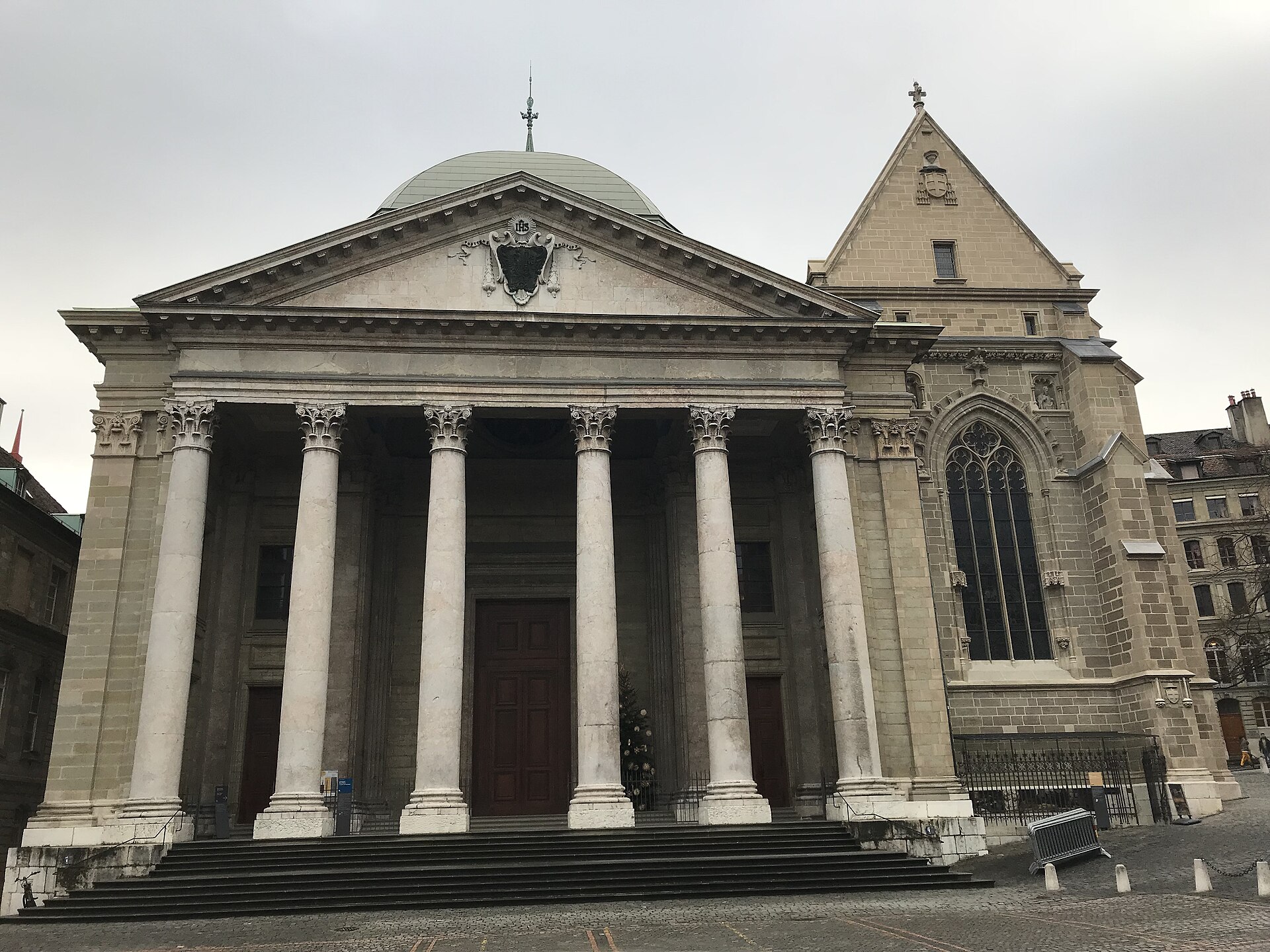 The columned neoclassical portico of St Pierre Cathedral in Geneva under a grey sky, with the Gothic Chapel of the Maccabees beside it